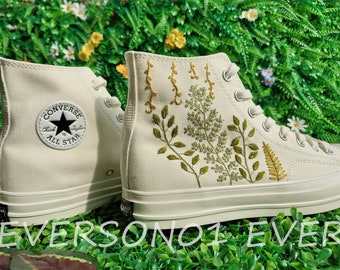 May include: White high-top Converse sneakers with green and yellow embroidered floral designs. The sneakers have a black Converse logo and the text "Chuck Taylor All Star" on the side.