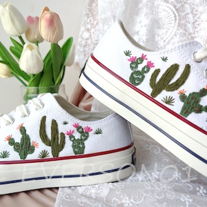 Puede incluir: Zapatillas bajas de lona blancas con diseños de cactus bordados en tonos verdes, rosas y naranjas. Las zapatillas tienen un ribete rojo y azul alrededor de la suela.