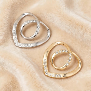 Pode incluir: Duas broches em forma de coração. Uma é prateada, a outra dourada. Ambas têm uma forma de coração menor dentro da maior. Cada broche é adornado com strass transparentes e brilhantes. O fundo é um tecido bege macio.