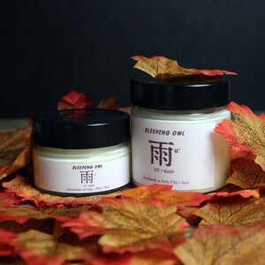 Op de afbeelding: Twee glazen kaarsen met zwarte deksels en witte etiketten. De etiketten hebben de tekst "Sleeping Owl" en een gestileerd Chinees karakter voor "regen". De kleinere pot is gelabeld met "90g (3 oz)" en de grotere pot met "170g (6 oz)". De potten zijn omringd door herfstbladeren.