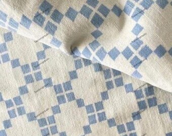 May include: Close-up of a cream-colored fabric with a repeating pattern of light blue diamond shapes. The fabric is folded, showing texture and depth. The design is geometric and modern, suitable for home decor.
