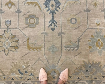 May include: A beige and grey patterned rug with floral and geometric designs. The rug features a central medallion and border with accents of blue and yellow. A pair of pink shoes and blue jeans are visible at the bottom of the image.