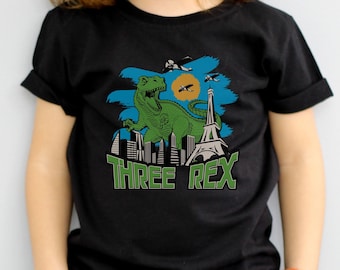 May include: A black t-shirt with a green dinosaur standing in front of a cityscape with the Eiffel Tower. The shirt has the text "THREE REX" printed on it.