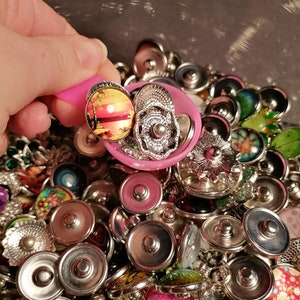 Peut inclure: Une cuillère rose remplie de boutons-pression de différentes couleurs et argentés. Les boutons présentent différents motifs, notamment floraux, géométriques et abstraits.