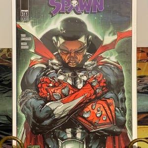 May include: Comic book cover for Spawn #311, featuring a portrait of a person with crossed arms, wearing a costume with red and black accents. The cover includes the text "In Memory of Chadwick Boseman (1976-2020)" and the Spawn logo.