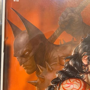 May include: Comic book cover for Wonder Woman #15, featuring a dark, stylized Batman figure with a spiked shoulder. The cover has an orange background with the title "Absolute Wonder Woman" at the top.