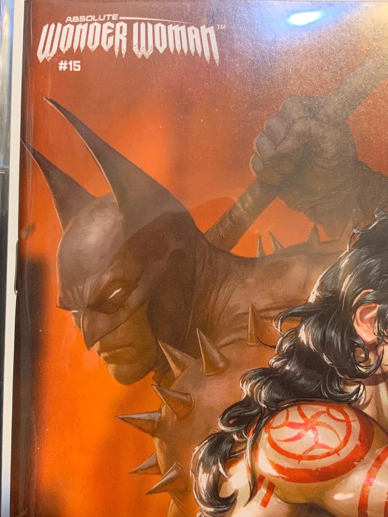 May include: Comic book cover for Wonder Woman #15, featuring a dark, stylized Batman figure with a spiked shoulder. The cover has an orange background with the title "Absolute Wonder Woman" at the top.