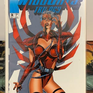 May include: Comic book cover for WildC.A.T.S. Trilogy #2, featuring a female character in a red bodysuit with a futuristic design. The cover has the title "WildC.A.T.S. Trilogy" in blue and the issue number and price are visible.