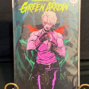 May include: A comic book titled Absolute Green Arrow 1 featuring a muscular character with short blonde hair wearing a pink tank top, black gloves, and black pants. The cover art displays a vibrant green and dark background with red accents.