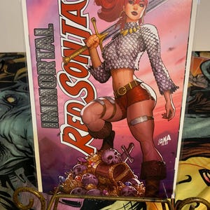 Może przedstawiać: Okładka komiksu przedstawiająca rudowłosą kobietę w zbroi kolczugowej i czerwonych spodenkach trzymającą miecz. Okładka nosi tytuł "Immortal Red Sonja" i numer "01". Kobieta stoi na stercie czaszek i skarbów.