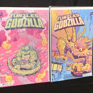 May include: Two Teenage Mutant Ninja Turtles vs. Godzilla comic books in protective sleeves. The first issue features a pink background with a turtle in a Godzilla-like pose. The second issue shows a yellow Godzilla-like creature in a city.