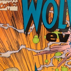 Pu&ograve; includere: Copertina di fumetto con il logo Marvel Comics e la parola "WOLVERINE" in grandi lettere blu. Lo sfondo &egrave; arancione con linee nere che si irradiano verso l'esterno ed elementi grafici verdi e bianchi.