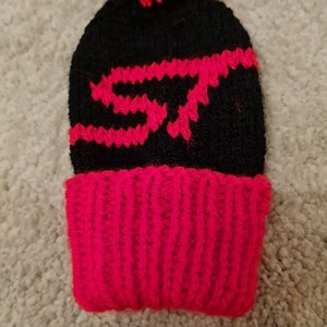 May include: Black and red knitted beanie with a pom pom on top. The beanie has a red stripe at the bottom and a black and red knit design on the top.