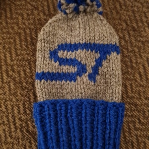 May include: A grey and blue knitted beanie hat with a blue pom-pom. The hat has a blue design on the front and a blue ribbed brim. This winter hat is ideal for keeping warm in cold weather.