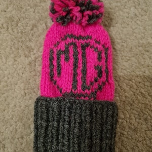May include: A pink knitted beanie with a grey pom pom and a grey knitted cuff. The beanie has a grey knitted design on the front.
