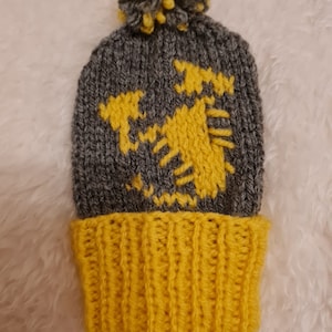May include: A grey and yellow knitted beanie with a pom pom on top. The beanie has a yellow knitted design on the front.