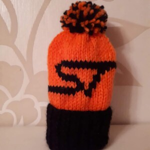 May include: Orange and black knitted hat with a pom pom on top. The hat has a black knitted design on the front.