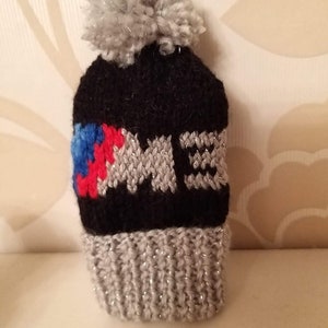 May include: A small, black knitted hat with a silver pom-pom. The hat has a silver knitted band with the letters "M E" and a colourful design of blue, red, and white. The bottom band is grey with silver thread.