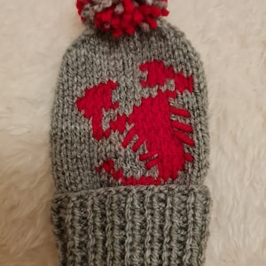 May include: Grey knitted beanie with a red pom pom and a red knitted design of a double-headed eagle on the front.
