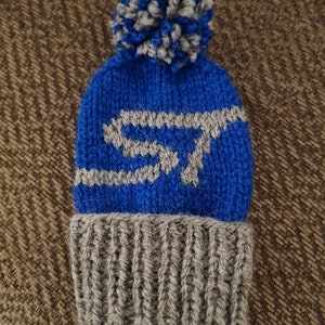 May include: A knitted beanie hat in blue and grey, with a pom-pom. The hat has a grey 'ST' design on the front. The lower section is grey, and the upper section is blue.