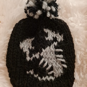 May include: Black knitted hat with a grey scorpion design and a black and white pom pom on top.