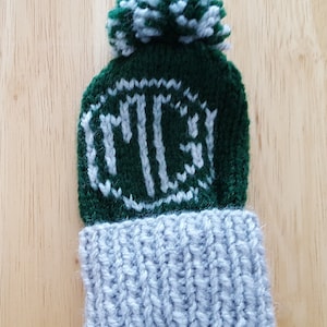 May include: A green knitted hat with a white pom pom and a white monogram "MCS" on the front. The hat has a grey knitted cuff.