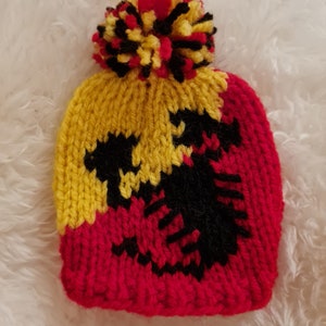 May include: A knitted hat with a red, yellow and black colour scheme. The hat has a pom-pom on top and a black dragon design.