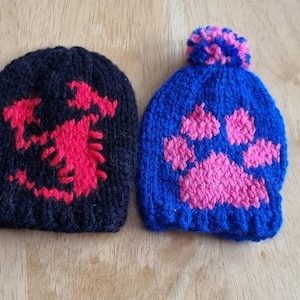 May include: Two knitted beanies. One is black with a red scorpion design. The other is blue with a pink paw print and a blue and pink bobble. The beanies are handmade.