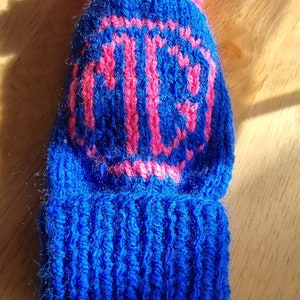 May include: A blue knitted hat with a pink pom-pom on top. The hat has a pink knitted design of the letters "P.C."