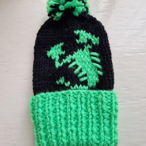 May include: Black knitted beanie with a green dragon design and a green pom pom on top.