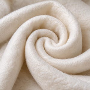 May include: A soft, off-white fabric with a dense, fuzzy texture arranged in a swirling, circular pattern. The material features a thick, plush pile that creates deep shadows within the folds, highlighting the uniform, light cream color throughout.