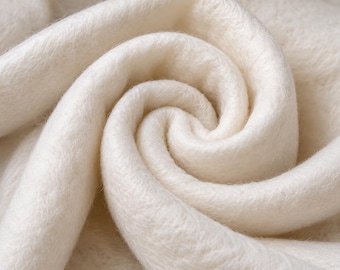 May include: A soft, off-white fabric with a dense, fuzzy texture arranged in a swirling, circular pattern. The material features a thick, plush pile that creates deep shadows within the folds, highlighting the uniform, light cream color throughout.