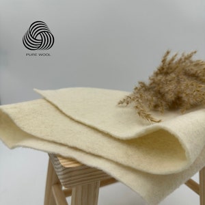 Può includere: Due quadrati di feltro di lana color crema impilati su uno sgabello di legno. Un filo d'erba secca è posto sopra il feltro. Il testo "PURE WOOL" è visibile nell'angolo in alto a sinistra dell'immagine.