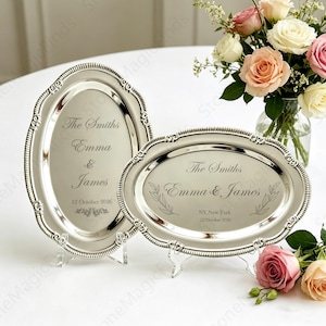 May include: Two silver-colored, oval trays with ornate edges, displayed on clear stands. The trays are engraved with the names "Emma & James" and the date "12 October 2026" and "NY, New York 12 October 2026". A bouquet of roses is in the background.