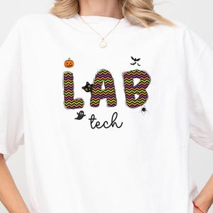 May include: White t-shirt with the text "LAB tech" in a colorful, Halloween-themed design. The letters are filled with a zig-zag pattern, and the design includes a pumpkin, bats, a ghost, and a spider.