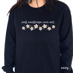 May include: Navy blue long-sleeved sweatshirt with the text "post anesthesia care unit" and a row of white daisies.