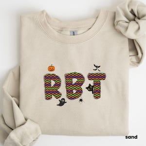 May include: A tan sweatshirt featuring a Halloween-themed design. The letters "RBT" are in a chevron pattern with orange, green, and purple colours. A black cat, a ghost, and a spider are also included in the design.