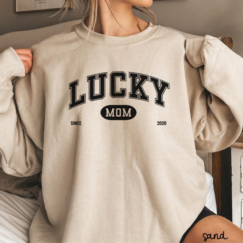 以下が含まれることがあります： ベージュのトレーナーに、黒い文字で「LUCKY MOM SINCE 2020」と書いてあります。文字は大学のフォントスタイルです。