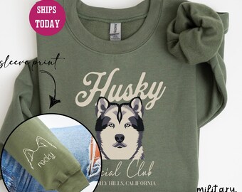 Op de afbeelding: Een olijfgroen sweatshirt met een witte husky-hond afbeelding en de tekst "Husky Social Club Beverly Hills, Californië". De linkermouw heeft een witte omtrek van een husky-hoofd met de tekst "rocky" eronder.