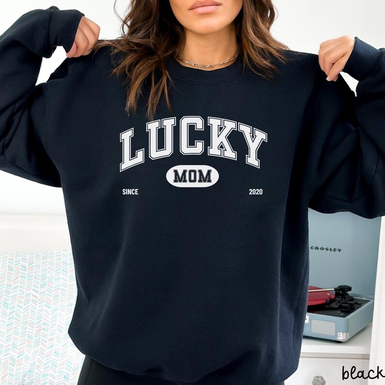 以下が含まれることがあります： 白い文字で「LUCKY MOM SINCE 2020」と書かれた黒いスウェットシャツ。テキストは大学風のフォントで、「MOM」の周りは白い楕円形になっています。