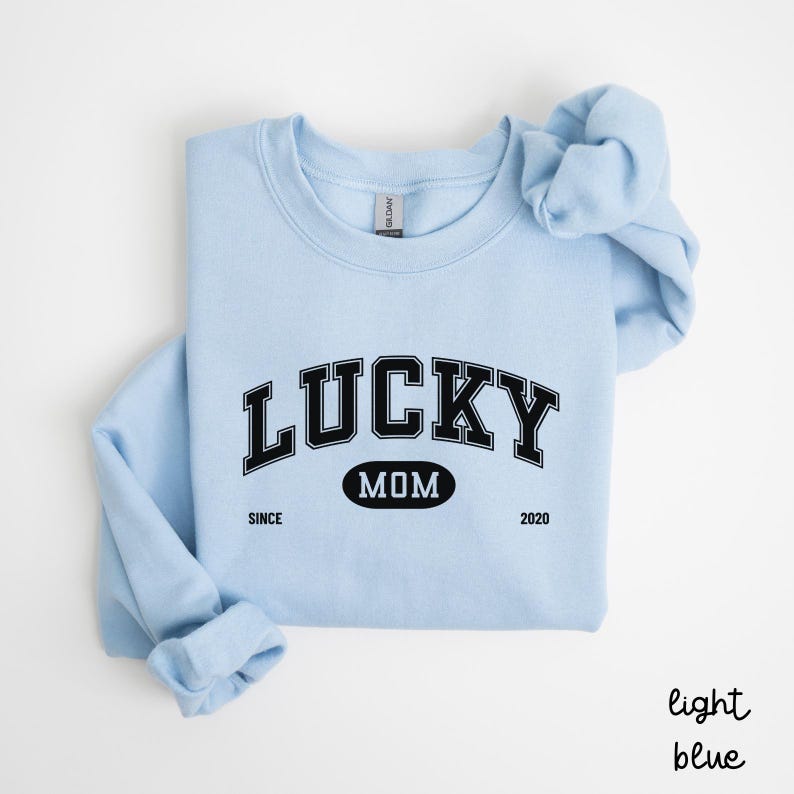 以下が含まれることがあります： 薄い青色のスウェットシャツに、黒い文字で「LUCKY MOM SINCE 2020」と書いてあります。