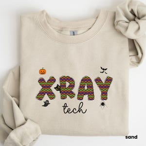 Puede incluir: Una sudadera de color arena con un diseño de temática de Halloween. El texto "XRAY tech" está escrito en negro con un patrón de zigzag en verde, naranja y morado. Hay pequeñas imágenes de una calabaza, un fantasma, una araña y murciélagos.