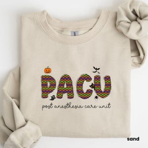May include: A tan sweatshirt with a Halloween-themed design. The word "PACU" is spelled out in black, green, and purple letters with a chevron pattern. The letters are surrounded by Halloween-themed graphics, including a pumpkin, a ghost, a spider, and bats. The text "post anaesthesia care unit" is printed below the design.