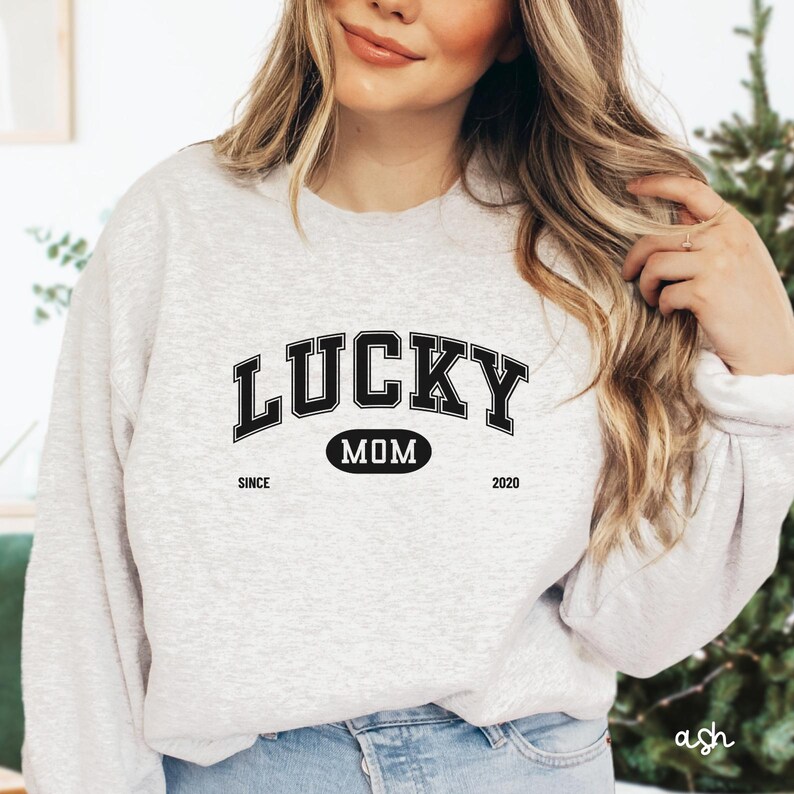 以下が含まれることがあります： 黒い文字で「LUCKY MOM SINCE 2020」と書かれた灰色のスウェットシャツ。 「MOM」という単語は黒い楕円形の中にあります。