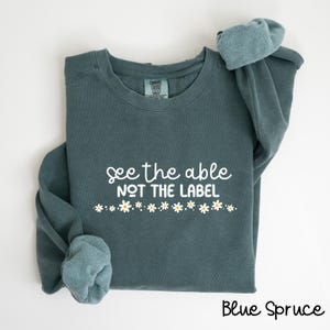 Może przedstawiać: Turkusowa bluza z napisem "See the able not the label" w białym kolorze. Pod napisem znajduje się rząd białych stokrotek z żółtymi środkami. Bluza jest złożona na pół, a widoczna jest metka z nazwą marki.