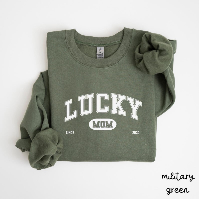 以下が含まれることがあります： 白い文字で「LUCKY MOM SINCE 2020」と書かれた緑色のスウェットシャツ。 「LUCKY」という文字は、大きく白いアーチ型のフォントで書かれています。「MOM」という文字は、白い楕円形の中に書かれています。