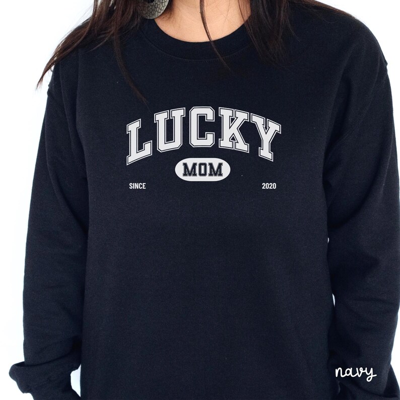 以下が含まれることがあります： 白い文字で「LUCKY MOM SINCE 2020」と書かれた黒い長袖スウェットシャツ。白い楕円形の中に黒い文字で「MOM」と書かれている。