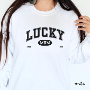以下が含まれることがあります： 白いスウェットシャツに、黒い文字で「Lucky Mom Since 2020」と書いてあります。