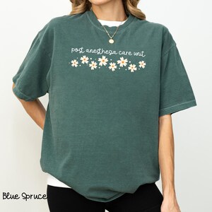 Könnte beinhalten: Ein blaugrünes T-Shirt mit dem Text "post anesthesia care unit" und einer Reihe weißer Gänseblümchen mit gelben Zentren.