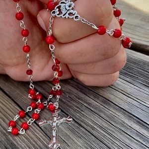 May include: A red rosary with a silver crucifix and a silver ornate centerpiece. The rosary is held in a hand.